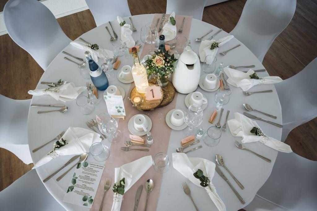 Gedeckter runder Hochzeitstisch im Eventhaus Boger – elegante Tischdekoration mit weißen Hussen, floraler Mitte und fein arrangiertem Besteck.