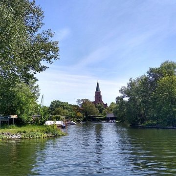 Blick vom Wasser auf die grüne Uferlandschaft in Brandenburg an der Havel mit einer Kirchturmspitze im Hintergrund.