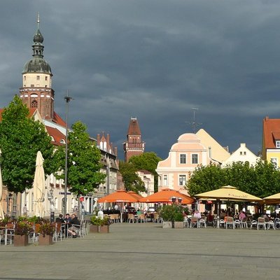 Großer Marktplatz in Cottbus mit historischen Bürgerhäusern, Sonnenschirmen und dem markanten Kirchturm im Hintergrund.