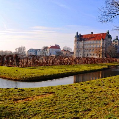 Das helle Renaissanceschloss Güstrow hinter einer grünen Wiese und einem Wassergraben an einem sonnigen Tag.
