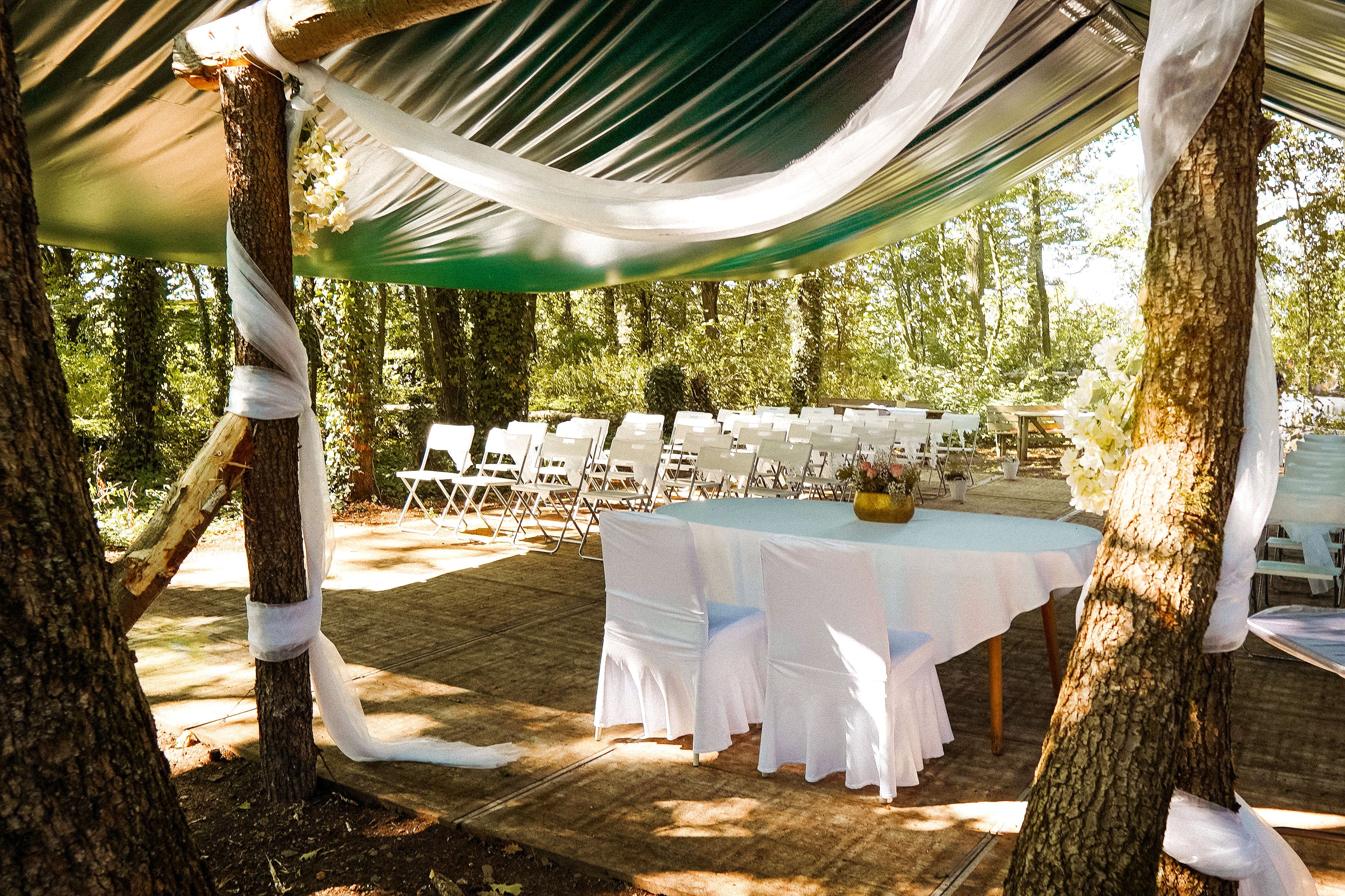 Blick durch den Traubogen auf den gedeckten Tisch und die weißen Stühle für die freie Trauung unter Bäumen nahe Dortmund