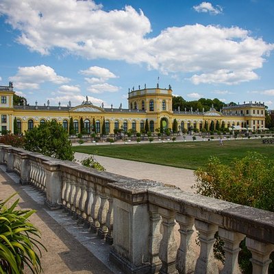 Das langgestreckte Schlossgebäude der Orangerie Kassel mit einer Steingalerie im Vordergrund unter blauem Wolkenhimmel.