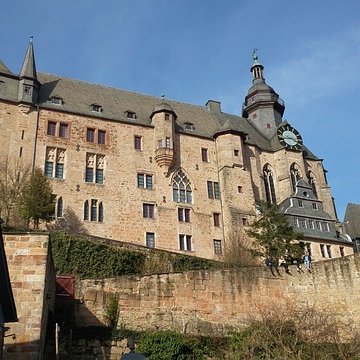 Blick hinauf zum monumentalen Marburger Schloss mit alten Festungsmauern und Fachwerk im Vordergrund unter blauem Himmel.