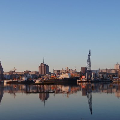 Blick auf den Rostocker Stadthafen bei spiegelglattem Wasser und blauem Himmel mit der Silhouette der Hansestadt.