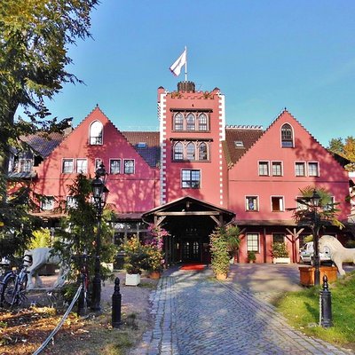 Die rote, schlossartige Fassade des Burghotels in Strausberg mit Zinnen, Turm und gepflastertem Eingangsbereich an einem sonnigen Tag.