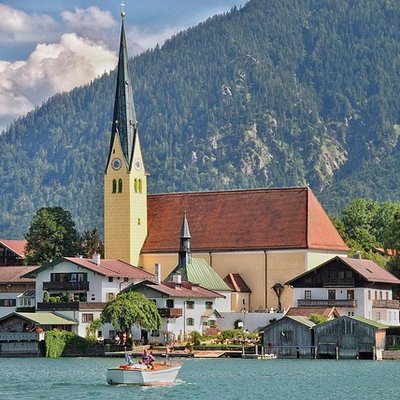 Blick über das türkisfarbene Wasser auf die markante Schlosskirche St. Quirinus am Tegernsee vor idyllischer Bergkulisse.