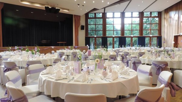 Festlich eingedeckter Hochzeitssaal im Kurhaus Bad Krozingen mit runden Tischen, weißer Tischdeko, lila Stuhlschleifen und großer Fensterfront mit Blick ins Grüne.