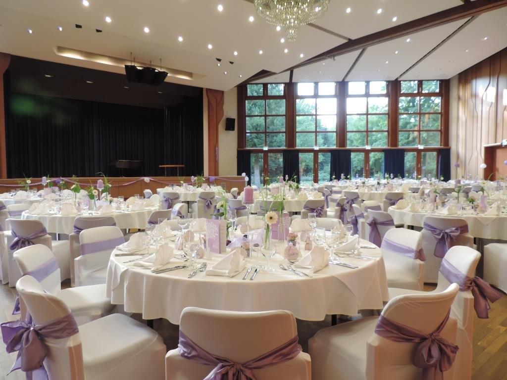 Festlich eingedeckter Hochzeitssaal im Kurhaus Bad Krozingen mit runden Tischen, weißer Tischdeko, lila Stuhlschleifen und großer Fensterfront mit Blick ins Grüne.