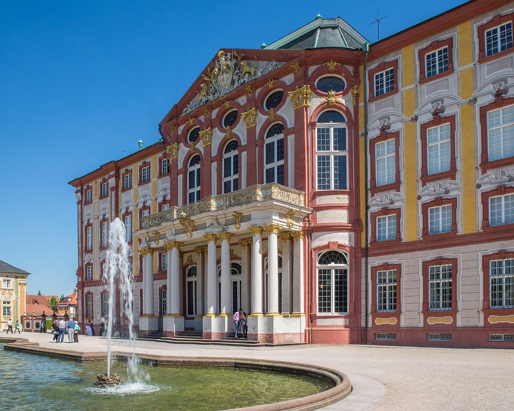 Die prachtvolle barocke Fassade von Schloss Bruchsal bei strahlendem Sonnenschein, im Vordergrund ein springender Springbrunnen.